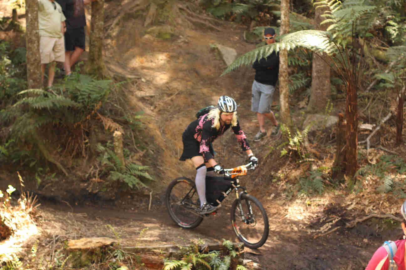 Right side view of a cyclist, wearing a dress and riding a Surly bike, on a dirt trail in a forest with people watching